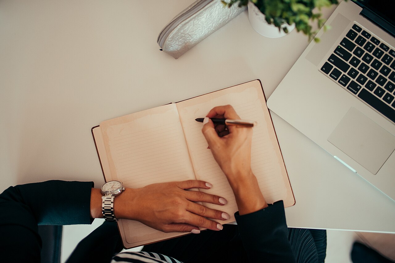 A person writing in a notebook next to an open laptop at a modern desk